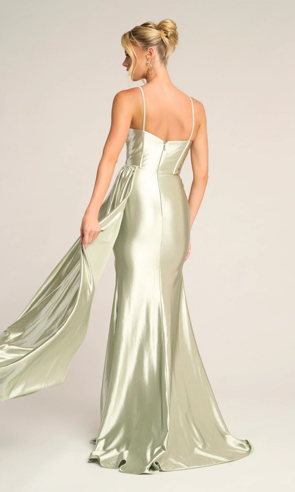 A woman in the Portia and Scarlett PS26270 long light green satin prom dress with thin straps stands facing away, holding part of the flowing fabric to the side.